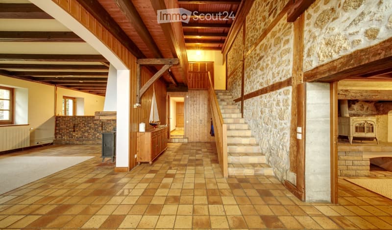 Spacious hallway with exposed wooden beams, stone walls, tiled floor, staircase leading upstairs, and a small cabinet with a decorative item on top.