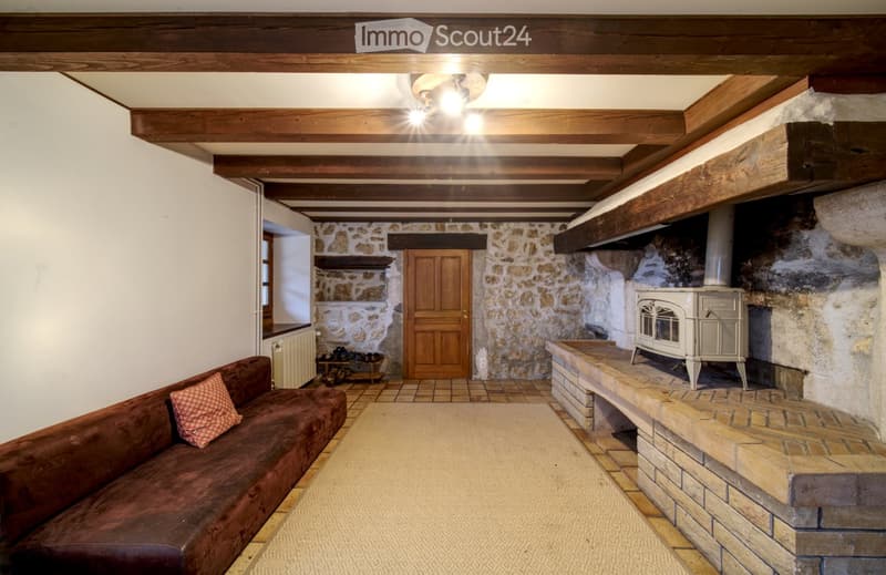 Living room with exposed wooden beams, stone walls, a brown leather sofa, a wooden stove, and a wooden door leading to another room.