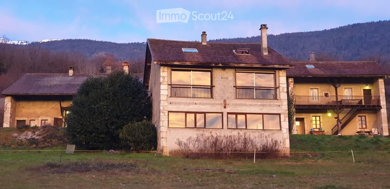 A two-story stone house with large windows and balconies, surrounded by a grassy area and trees, with mountains in the background.