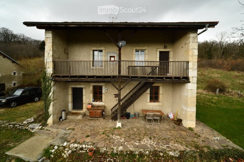 Two-story stone house with a wooden balcony, stone patio, and outdoor furniture including a table and chairs.