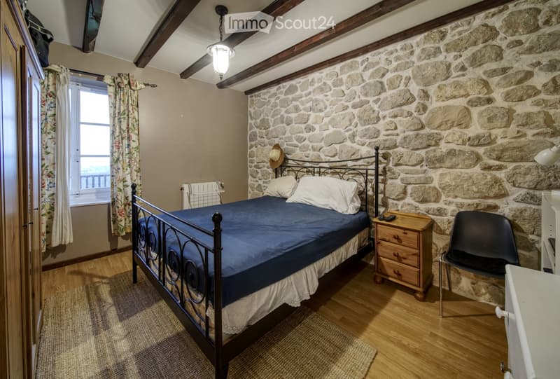 Cozy bedroom with exposed stone walls, wooden floor, and a metal-framed bed with blue bedding. Includes a wooden nightstand, a black chair, a window with floral curtains, and a hat on the wall.