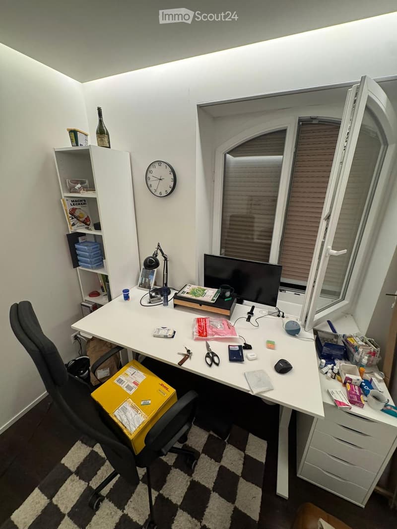 A well-organized home office with a white desk, ergonomic chair, computer monitor, desk lamp, and various office supplies. The room features a large window with blinds, a shelf with books and decorative items, and a patterned rug on the floor.