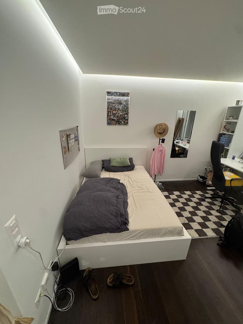 A small, modern bedroom with a single bed, a desk with a chair, a mirror, and a shelf. The room features a slanted ceiling and a checkered rug on the floor.