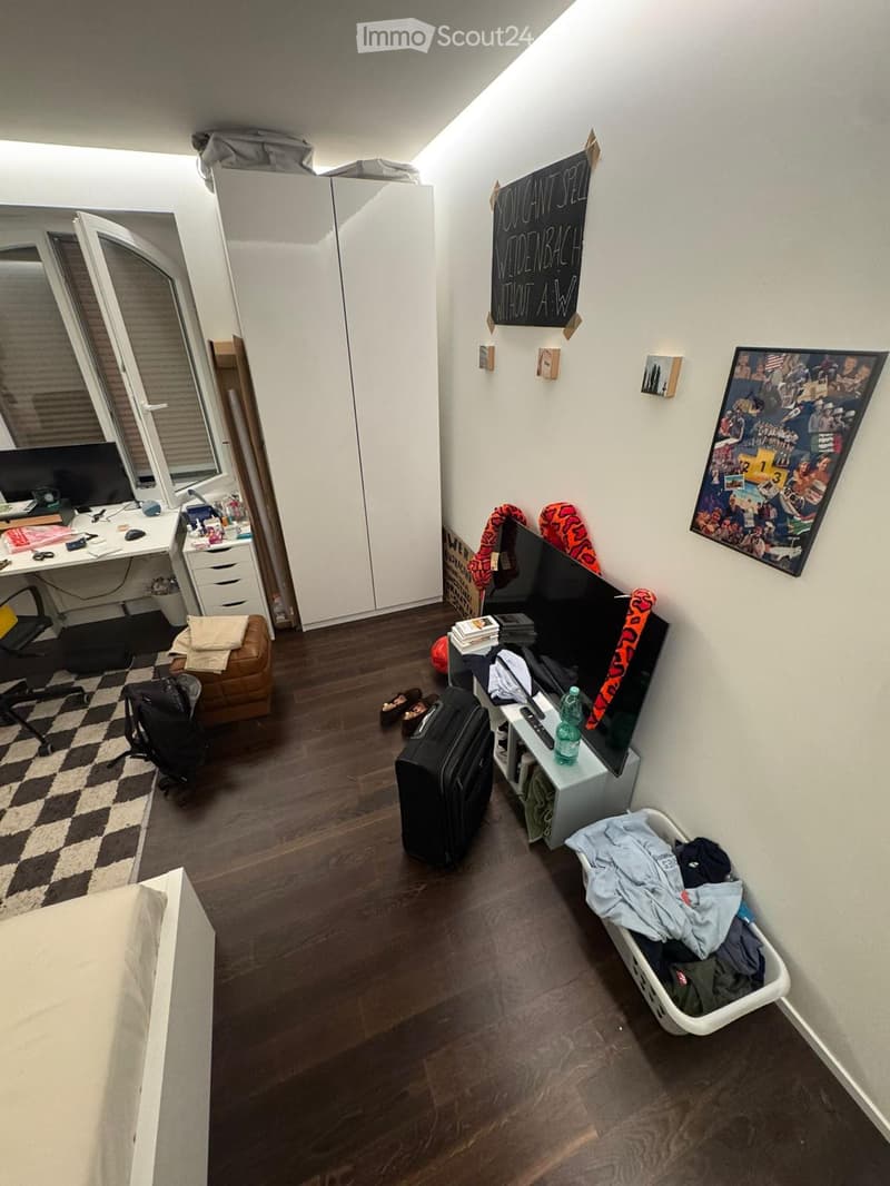 A furnished room with a bed, desk, chair, screen, table, stool, mirror, wardrobe, backpack, suitcase, laptop, remote control, bottle, picture frame, poster, and laundry basket.