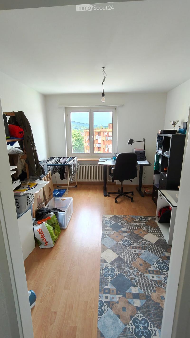 A furnished room with a wooden floor, a large window with a view, a desk with a chair, a lamp, a shelf, and a radiator.