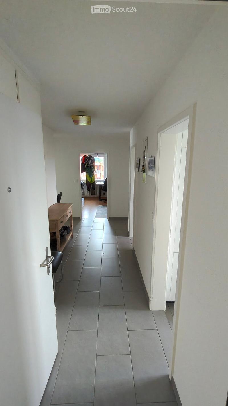 A long, narrow hallway with tiled flooring and white walls, featuring a small wooden shelf with shoes and a chair, and a partially open door leading to another room with visible furniture.