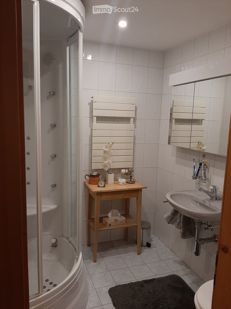 Modern bathroom with white tiles, a bathtub, a shower, a sink with a mirror, a small wooden table with a clock and flower, and a radiator.