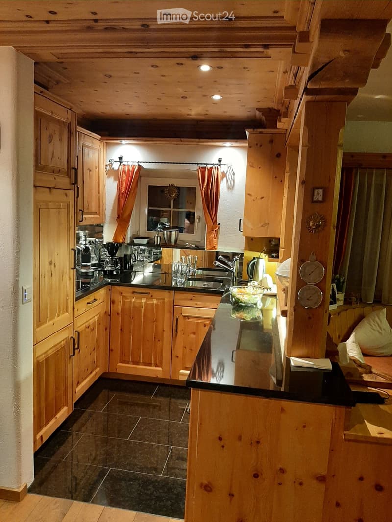 A cozy kitchen with wooden cabinets, a black countertop, and various appliances including a coffee maker, sink, and fridge. The space features a window with orange curtains and a small seating area visible in the background.