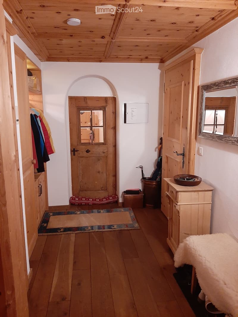 Wooden-floored hallway with white walls, wooden ceiling beams, and a small wooden cabinet with a mirror and bowl on top. A wooden door with glass panels leads to another room, and a small rug is placed near the door.