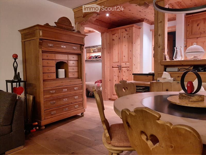 A cozy, wooden-furnished room with a sofa, round wooden table, chairs, tall wooden cabinet, and decorative items on shelves and surfaces.