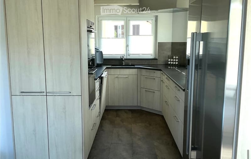 Modern kitchen with light wood cabinets, stainless steel appliances, and a dark countertop. Features a sink, faucet, and a window with a view.