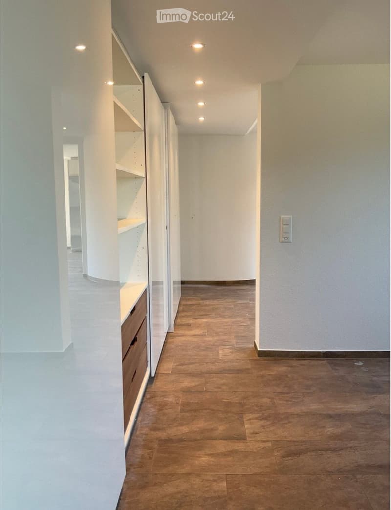 Modern hallway with wooden flooring, white walls, built-in shelving unit with drawers, recessed ceiling lights, and a power outlet on the wall.