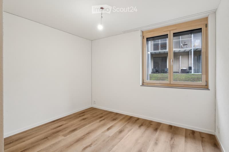 Empty room with light wooden flooring and white walls, featuring a large window with wooden frame and blinds, offering a view of an outdoor area with furniture.