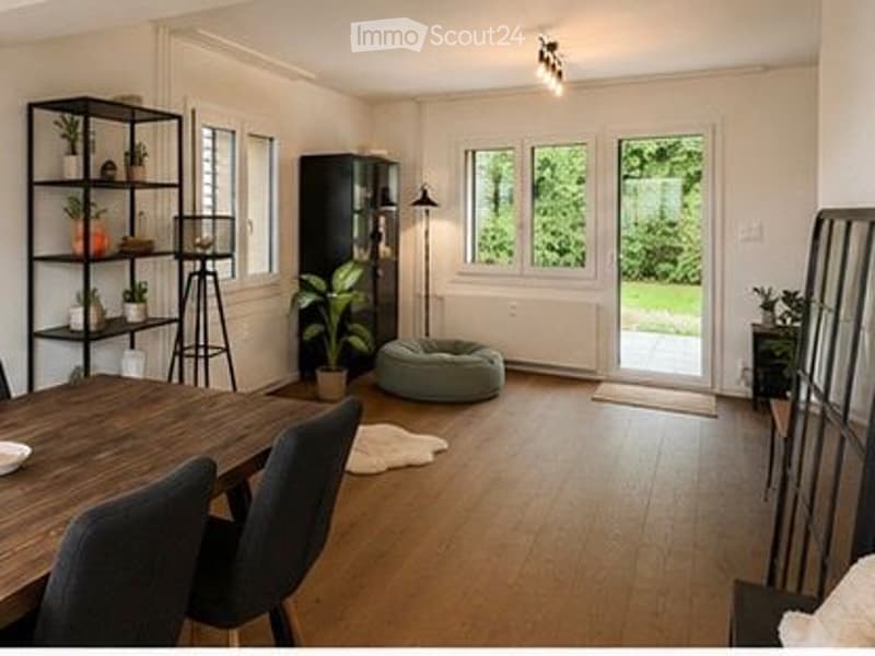 Bright, open-plan living and dining area with wooden flooring, large windows, a wooden dining table with chairs, a shelf with plants, a floor lamp, a dog bed, and a small rug.
