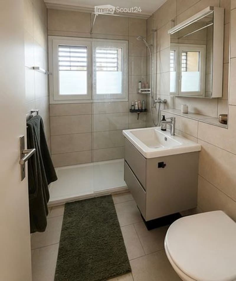 Modern bathroom with a glass-enclosed shower, a white sink with a mirror, a toilet, and a towel hanging on the wall. The space features beige tiles and a small window with blinds.