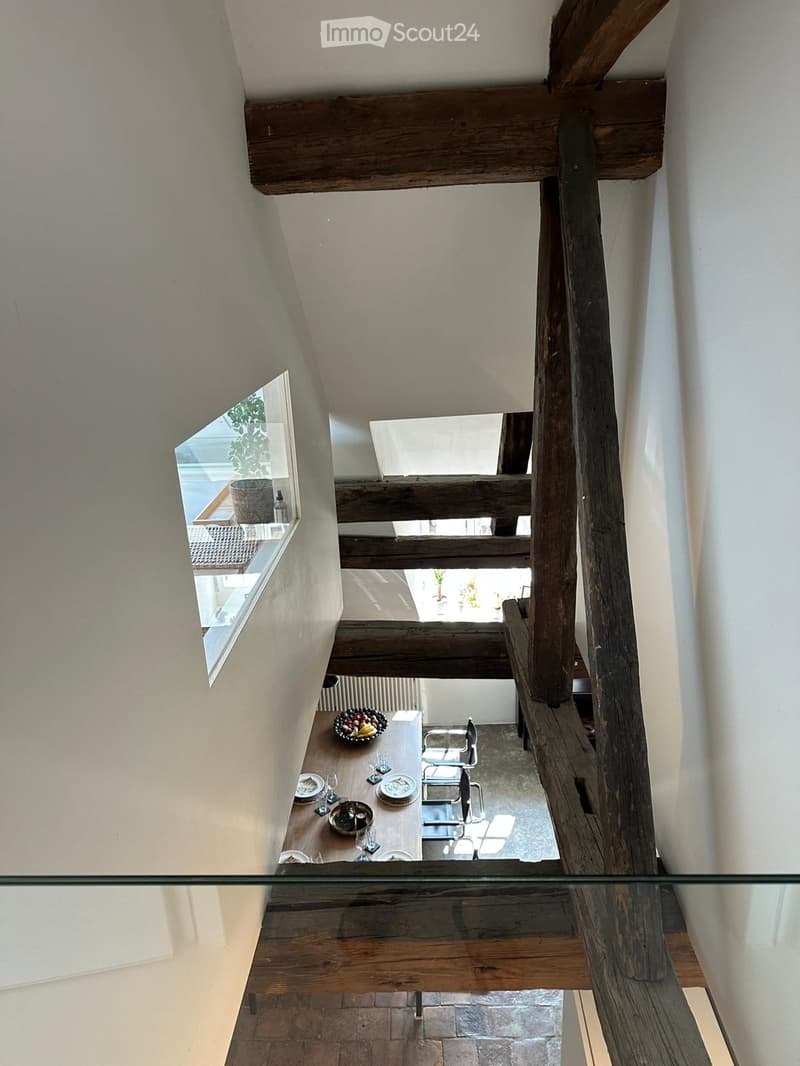 A modern, open-concept hallway with exposed wooden beams and a glass railing, leading to a dining area with a table and chairs.
