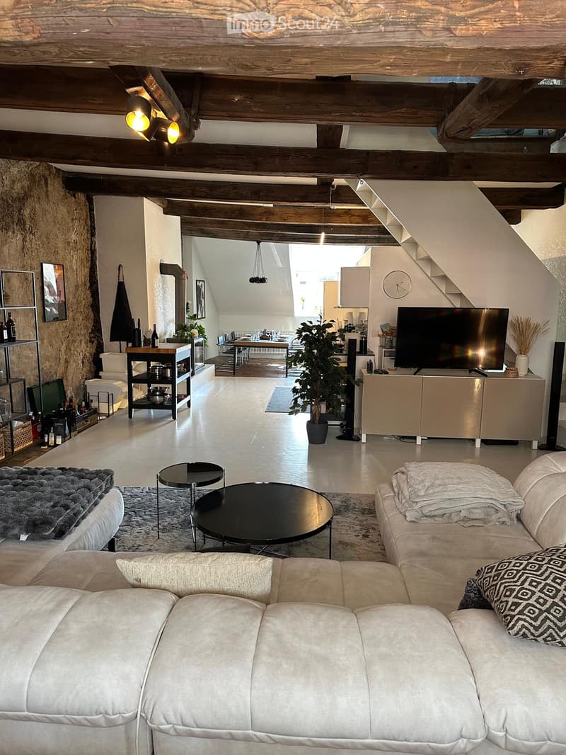 Modern living room with exposed wooden beams, a large white sectional sofa, a black coffee table, a flat-screen TV on a cabinet, and a dining area visible in the background.