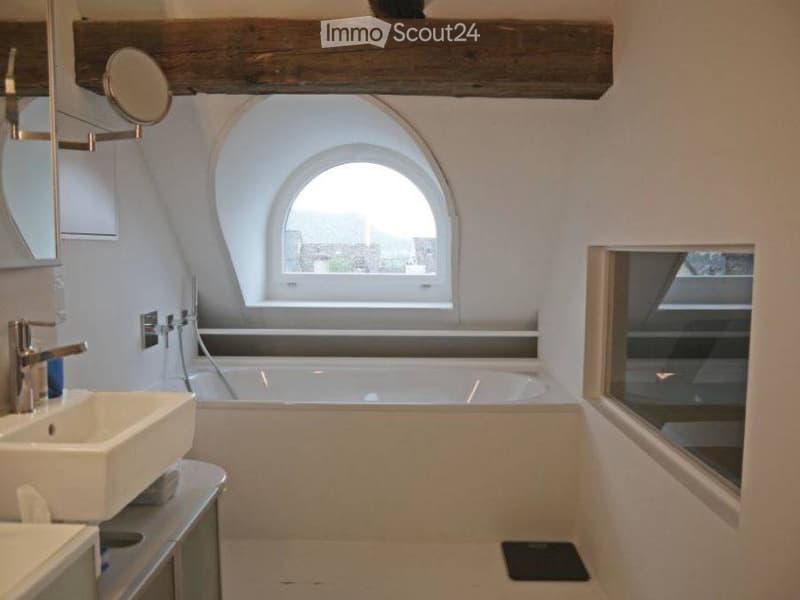 Cozy attic bathroom with exposed wooden beams, arched window, white sink, bathtub, and modern fixtures.