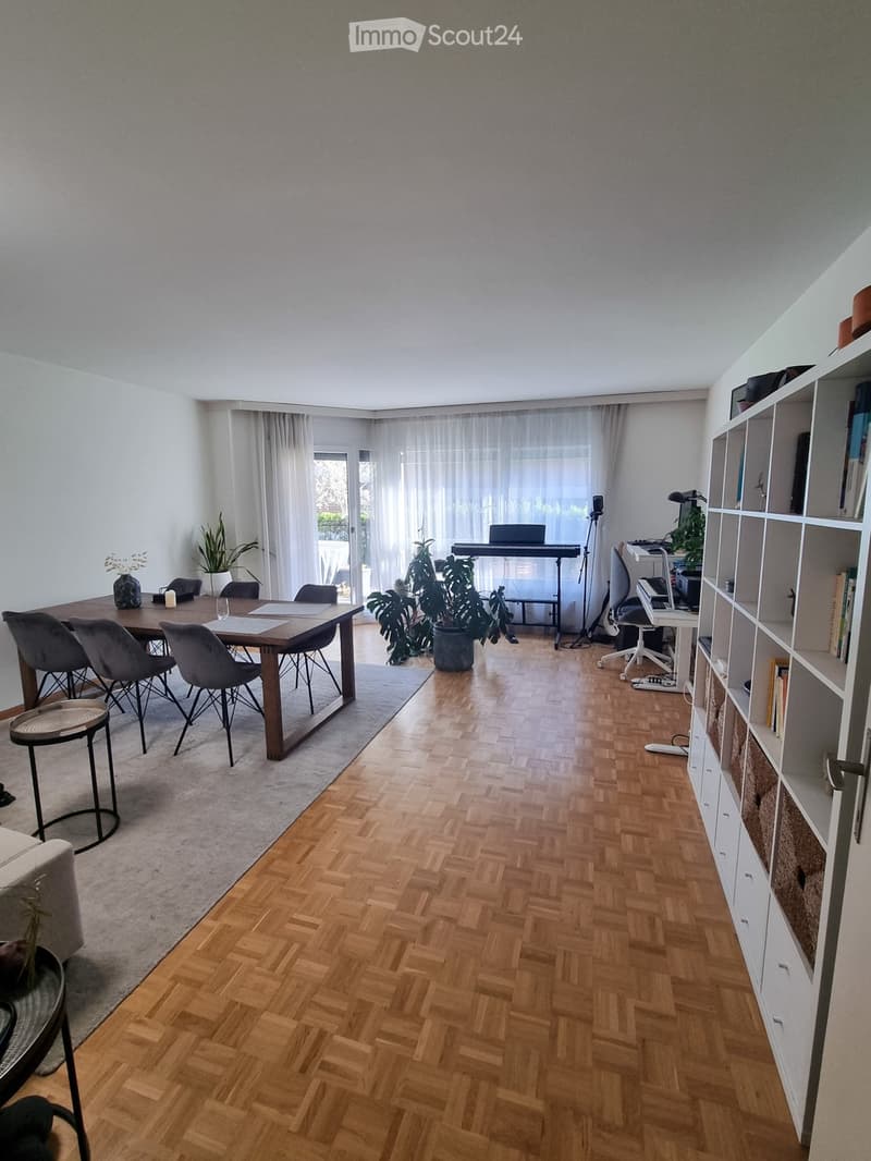 A modern, well-lit room with wooden herringbone flooring, a large window with sheer curtains, a dining area with a wooden table and chairs, a small sofa, a round stool, a bookshelf, a piano, and a desk with a chair.