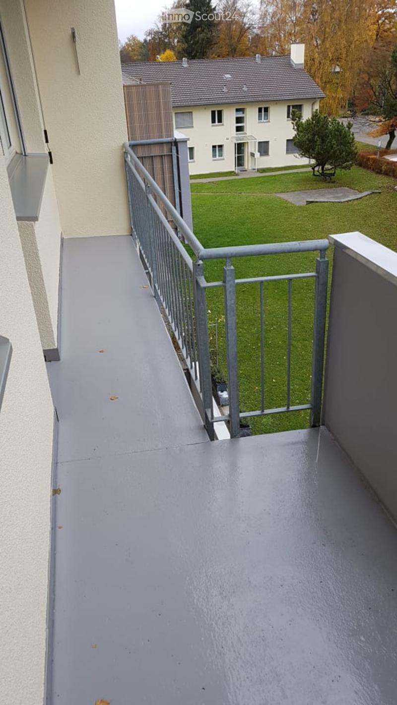 Balcony with grey floor and metal railing, overlooking a green lawn and neighboring house.