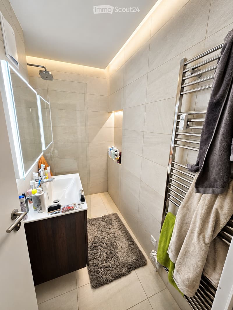 Modern bathroom with beige tiles, a dark wooden sink cabinet, a large mirror, a towel rack with towels, a shower area, and a fluffy rug on the floor.