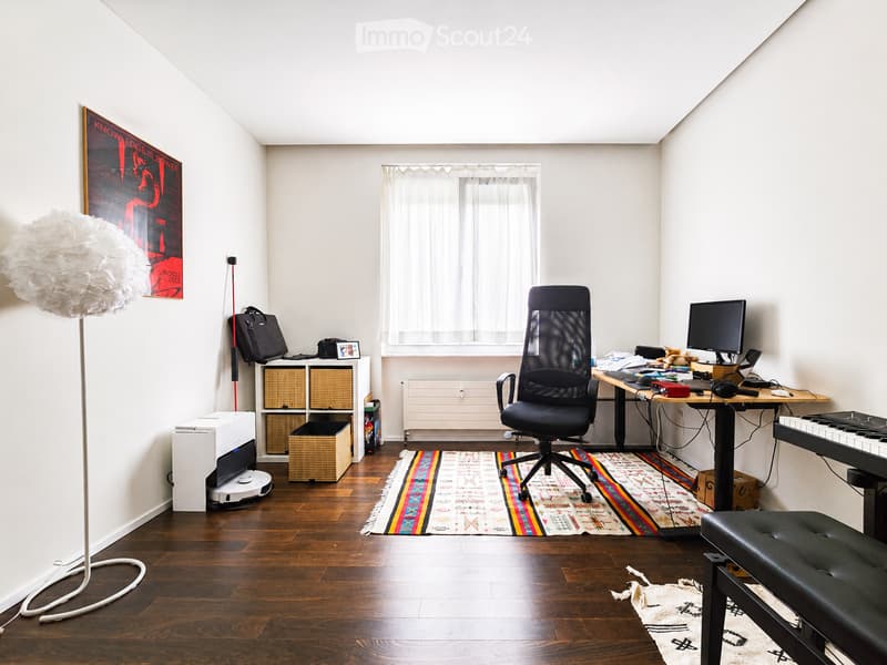 A modern, well-lit room with wooden flooring, a large window with white curtains, a black office chair, a desk with a computer, a floor lamp, a storage unit with woven baskets, a colorful rug, and a small piano bench.
