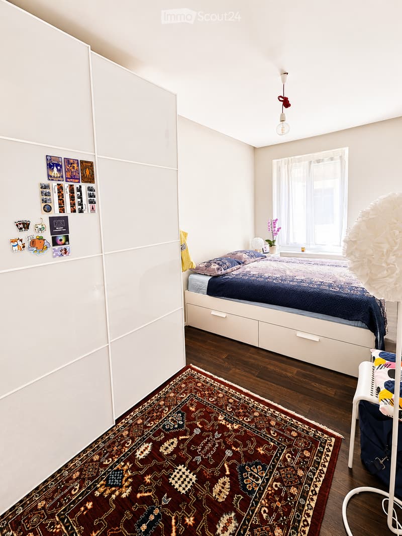 Cozy bedroom with a bed, pillows, a patterned rug, a chair, a hanging lamp, and a potted plant near the window.