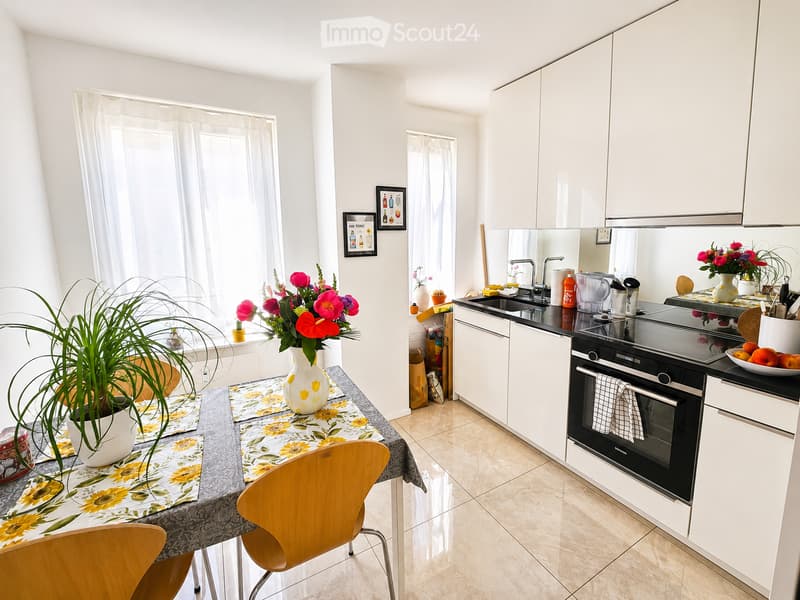 Bright kitchen with white cabinets, black countertops, and a dining area. Features a table with a floral tablecloth, chairs, a vase with flowers, and a potted plant on the windowsill.