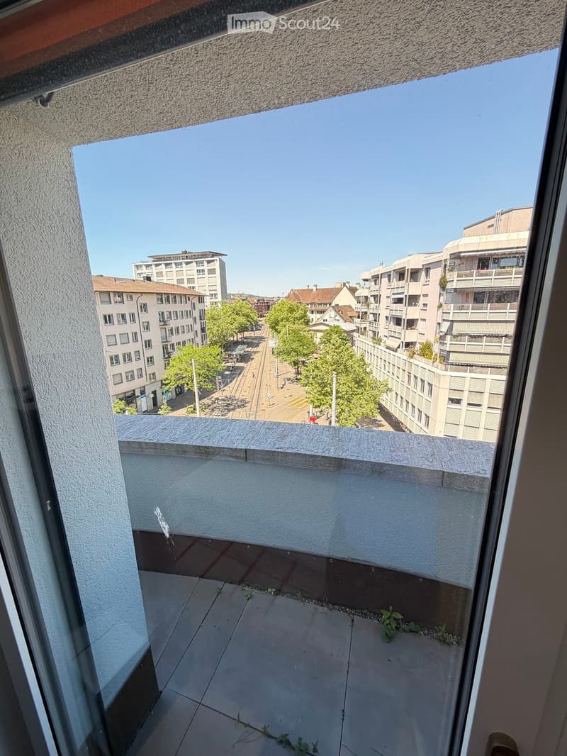 Balcony with tiled floor, overlooking a city street with buildings and trees.