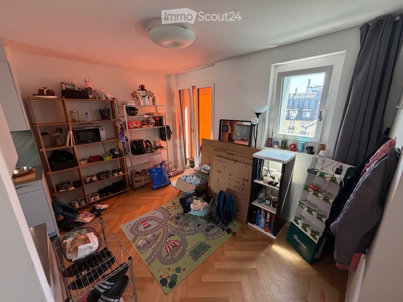 A well-lit room with wooden flooring, shelves filled with various items, a microwave, a refrigerator, a mirror, a lamp, a colorful play rug, and storage boxes.