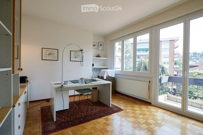 Bright, modern room with wooden flooring, large windows, and a balcony. Features a white desk, green chair, floor lamp, and picture frames on the wall.
