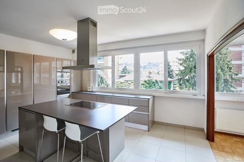Modern kitchen with a central island, built-in stove, sink, and large windows offering a view of greenery outside.