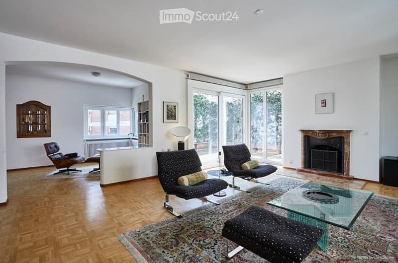 Modern living room with hardwood flooring, fireplace, large windows, and contemporary furniture including a sofa, coffee table, armchairs, and a floor lamp.