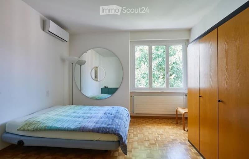 A modern, minimalist bedroom with a single bed, a large round mirror, a wooden wardrobe, a small stool, and a floor lamp. The room features light-colored walls, wooden flooring, and large windows with a view of greenery.