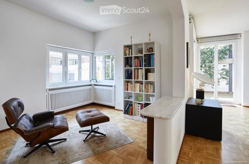 Bright, modern living room with large windows, wooden flooring, leather armchair, ottoman, bookshelf, and a small table with a lamp and cup.