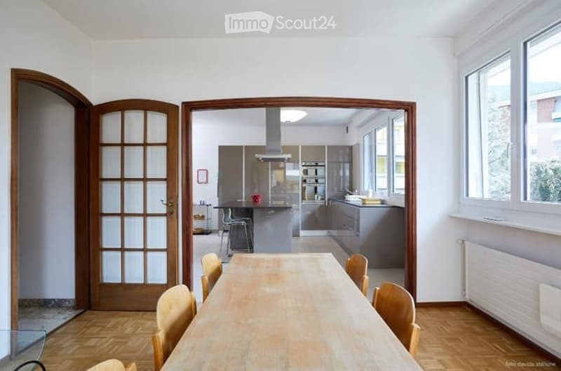 Open-plan dining and kitchen area with wooden flooring, large dining table surrounded by chairs, modern kitchen with stainless steel appliances, and ample natural light from large windows.