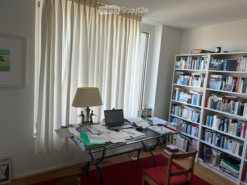 A well-furnished study or office space with a glass desk, laptop, bookshelves filled with books, a table lamp, and a wooden chair.
