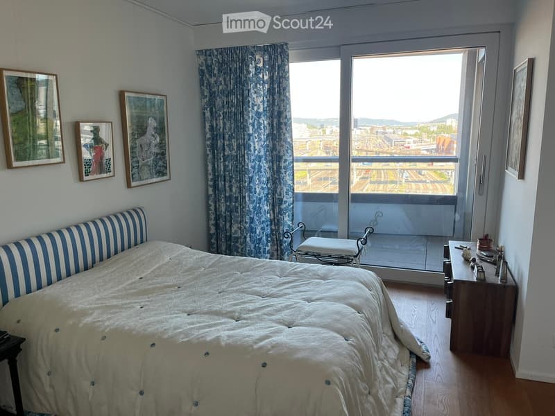 Bright bedroom with a large window offering a city view, featuring a bed with striped headboard, a nightstand, a chair, and several framed pictures on the wall.