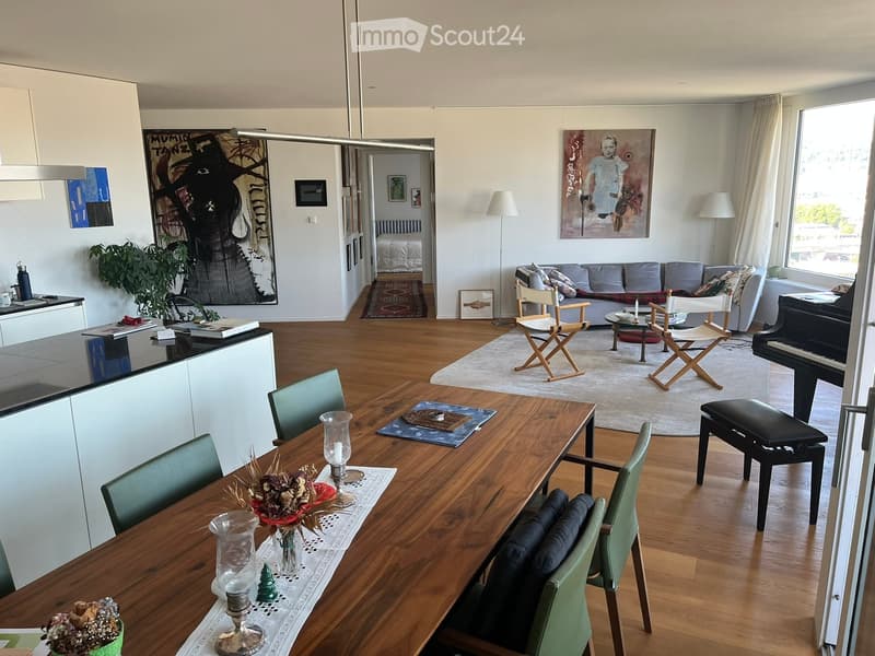 Open-plan living and dining area with wooden flooring, a grey sofa, a wooden dining table with green chairs, a black piano, and a modern kitchen area with white cabinets.