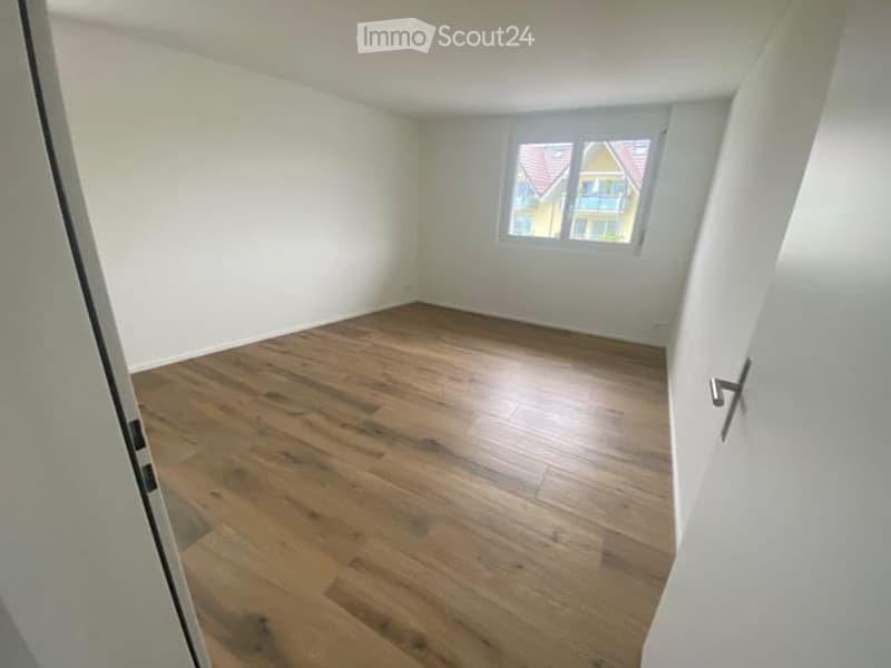 Empty room with wooden flooring and white walls, featuring a single window with a view of a neighboring building.