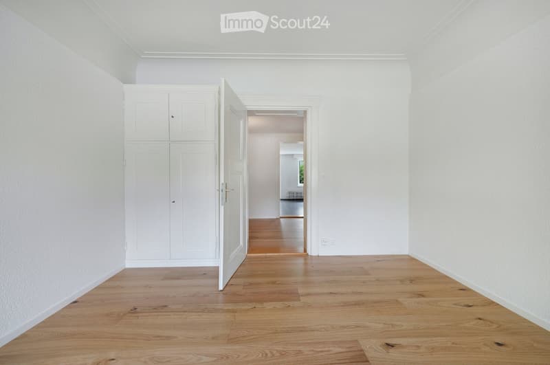 Empty hallway with light wooden flooring, white walls, and a built-in white cabinet on one side. The hallway leads to another room with visible furniture.