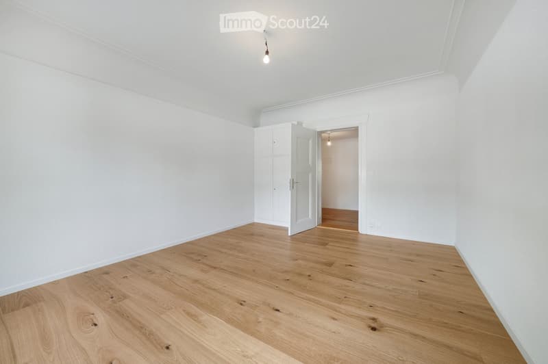 Empty room with light wooden flooring and white walls, featuring a single open door leading to another room.
