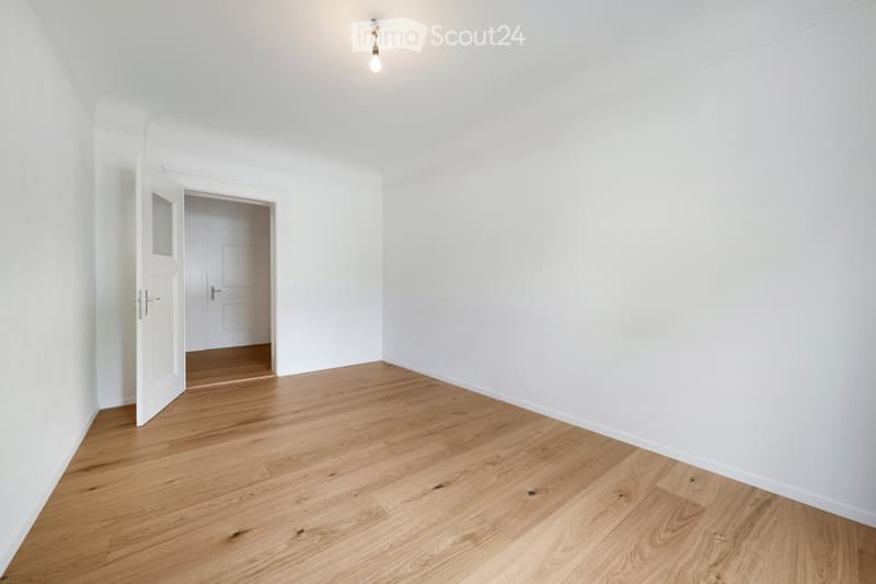 Empty room with light wooden flooring and white walls, featuring an open doorway leading to another room.