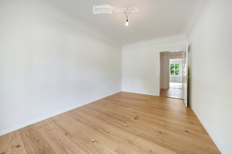 Empty hallway with light wooden flooring and white walls, featuring a single light fixture and an open doorway leading to another room.