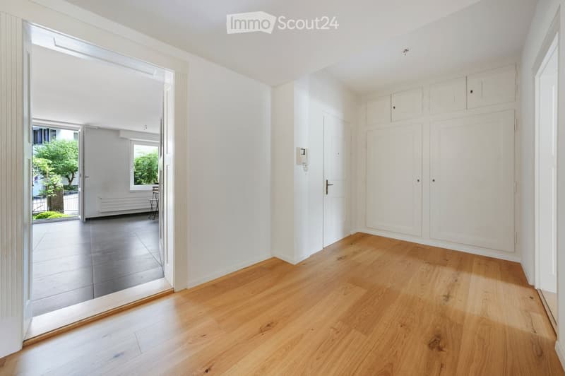 Bright, minimalist hallway with light wooden flooring, white walls, and built-in storage cabinets. An open doorway leads to another room with a tiled floor and a view of a green outdoor area.
