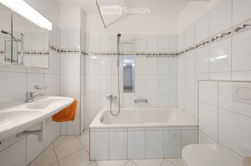 Modern bathroom with white tiles, a bathtub, a sink, a toilet, and a showerhead. The walls feature decorative border tiles, and there is a mirror above the sink.