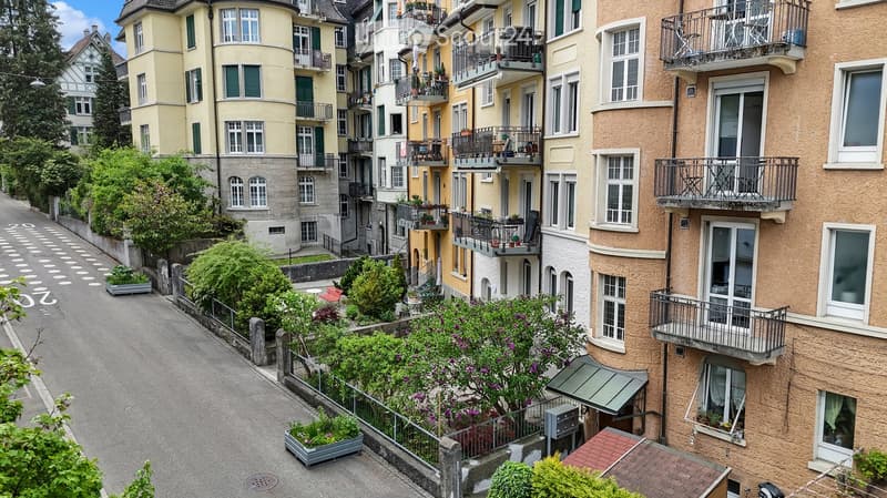 A picturesque street lined with multi-story residential buildings, featuring balconies, greenery, and colorful facades.