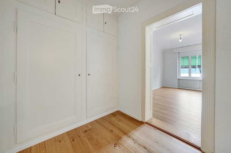 Empty hallway with wooden flooring, white walls, and a closed door leading to another room with large windows.