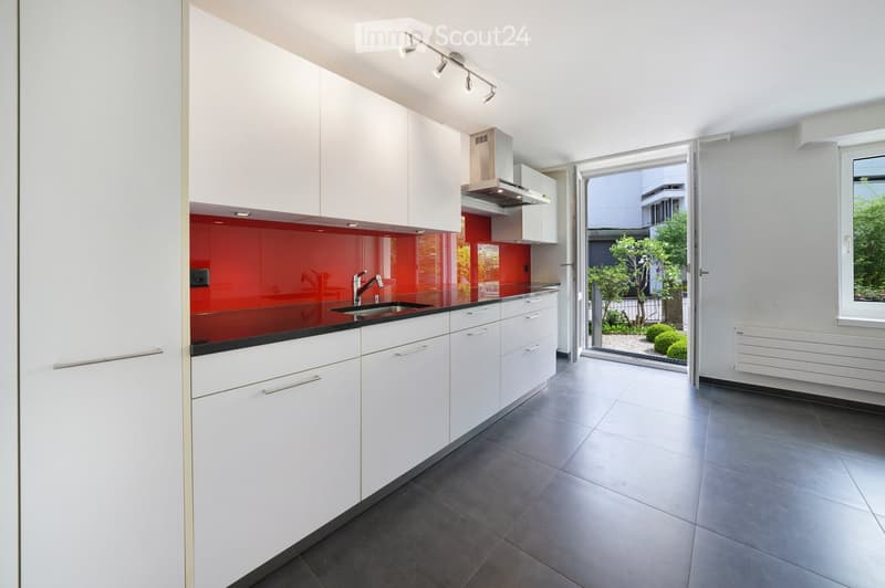 Modern kitchen with white cabinets, red backsplash, black countertop, stainless steel sink, and range hood. Open to a small outdoor area with greenery.