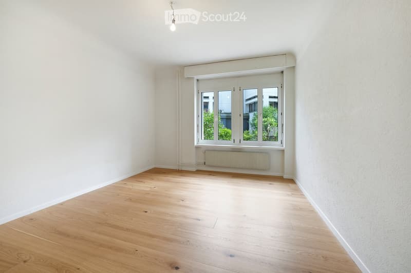 Empty room with light wooden flooring, white walls, and a large window with white frames. A radiator is visible below the window, and a single pendant light hangs from the ceiling.